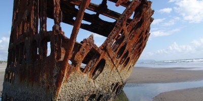 A color picture of a rusting shipwreck on the Oregon coast. The only things left are the orange rusting steel frame listing heavily to port on a sandy beach.