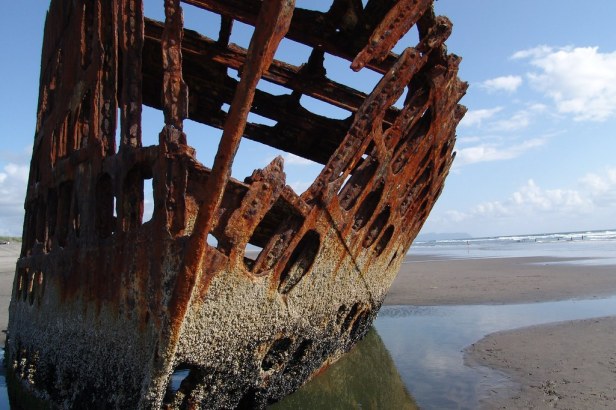 A color picture of a rusting shipwreck on the Oregon coast. The only things left are the orange rusting steel frame listing heavily to port on a sandy beach.