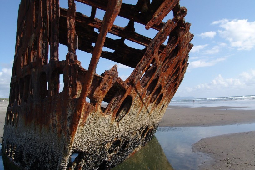 A color picture of a rusting shipwreck on the Oregon coast. The only things left are the orange rusting steel frame listing heavily to port on a sandy beach.