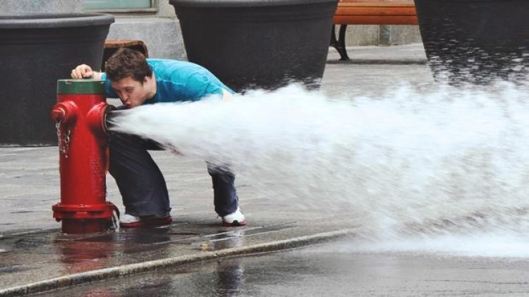 A man bending over the forceful stream exiting a fire hydrant attempting to take a drink from it.