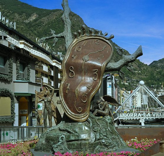 A sculpted version of Dali's Persistance of Memory painting. It depicts a "melting" clock face draped over the limb of a dead tree. The metal is dark. It is in an outdoor square with a mountain in the back and apartments to the left.